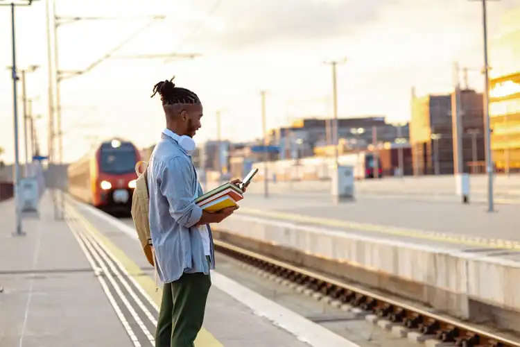 Homem carrega livros e utiliza smartphone numa estação de trem.