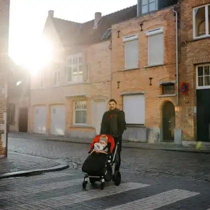 Pai segurando carrinho de criança na Bélgica, durante dia de sol e frio. Ambos estão agasalhados.