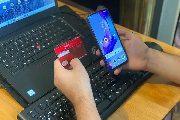 Pessoa segurando um cartão de crédito e um celular na mão na frente do computador