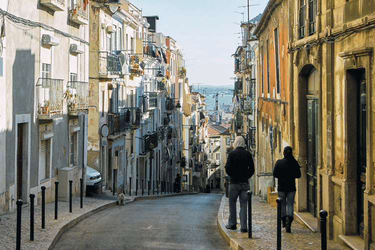 Uma rua estreita no centro de Lisboa, com casas coloridas e, ao fundo, o rio Tejo