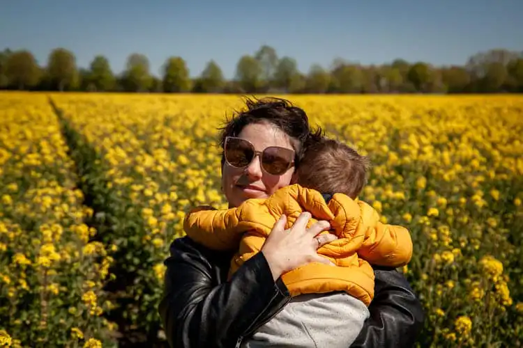 mulher de óculos escuros segura bebê nos braços em frente a um campo de flores