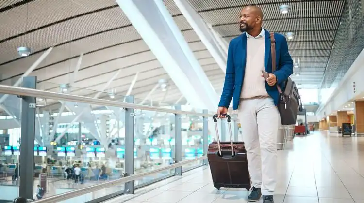 Homem caminhando no aeroporto com mala e passagem aérea para Londres executiva nas mãos.