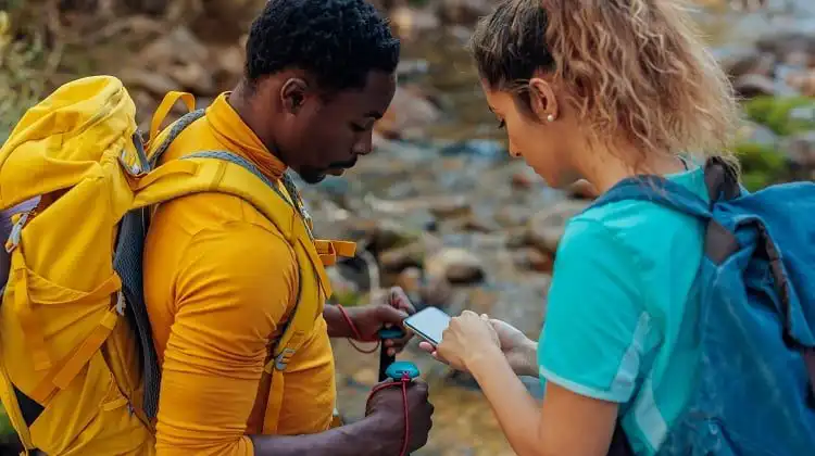 Homem negro e mulher morena de cabelos loiros olhando um celular, em meio a uma trilha