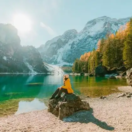 Mulher sentada em rocha, na frente de um lago. Ela usa roupas de frio e um casaco amarelo vibrante. Para viajar, seguro viagem vale a pena e pode ser obrigatório.
