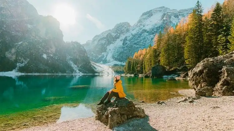 Mulher sentada em rocha, na frente de um lago. Ela usa roupas de frio e um casaco amarelo vibrante. Para viajar, seguro viagem vale a pena e pode ser obrigatório.