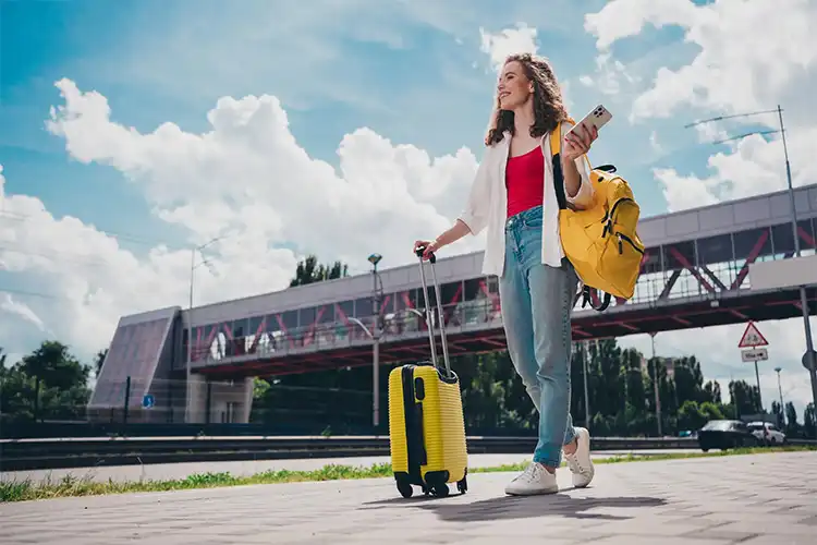 Utilizando um celular, jovem aguarda transporte com bagagens e celular em mãos.