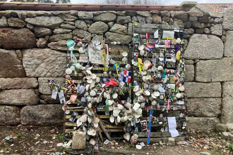 Cerca com centenas de vieiras e mensagens penduradas frente a um muro de pedra.