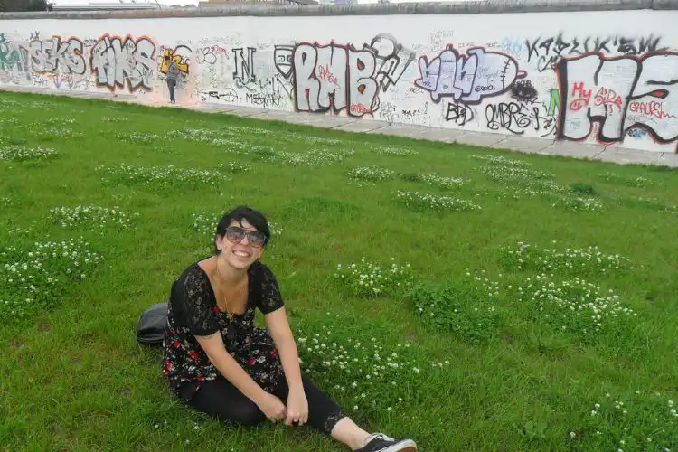 Jovem de óculos escuros sentada na grama em frente ao Muro de Berlim.