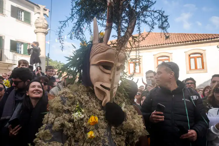 Pessoa com máscara de madeira e roupa de plantas em meio à multidão.