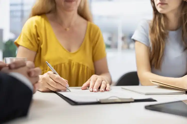 Mulher assina um contrato em uma mesa de reunião acompanhada por outras duas pessoas