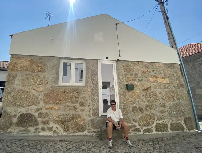 Mulher com cabelo curto e camiseta branca sentada no degrau da entrada de uma pequena casa de pedra.