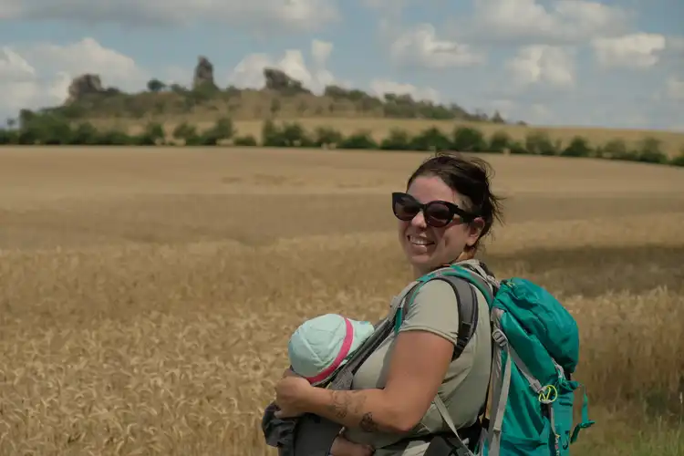 Mulher em frente a um campo seco sorri enquanto segura em bebê e carrega uma mochila verde.