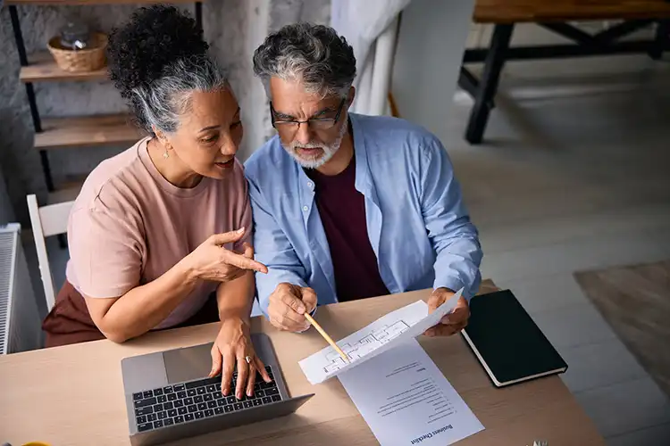 Casal sênior analisando planta de sua nova casa usando laptop