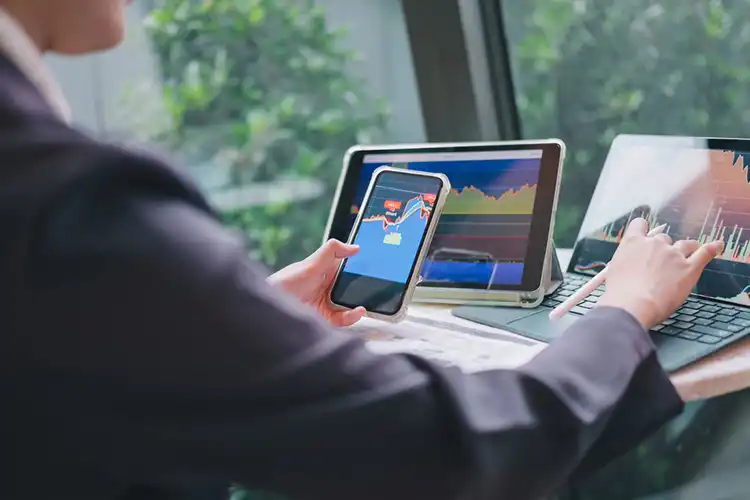 Jovem analisando gráficos de investimentos simultaneamente em programa no celular, tablet e notebook