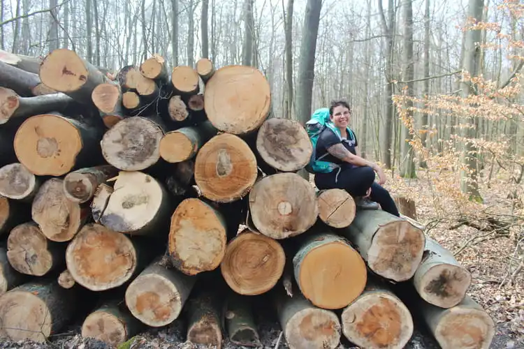 Mulher com mochila verde nas costas sentada em troncos cortados no meio de uma floresta na Alemanha.