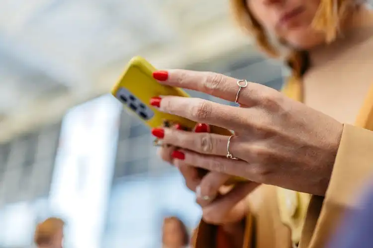 Mulher segurando um celular amarelo e digitando, com foco nas mãos e unhas vermelhas.