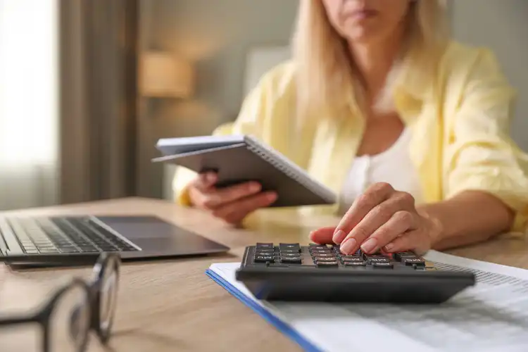 Pessoa usa calculadora e caderno, com laptop à mesa.