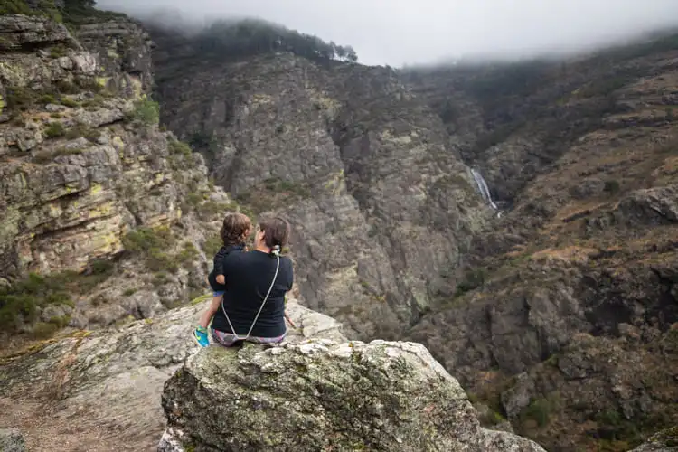 Mulher de costas com camisa preta e menina pequena no colo observa o abismo em paisagem rochosa.