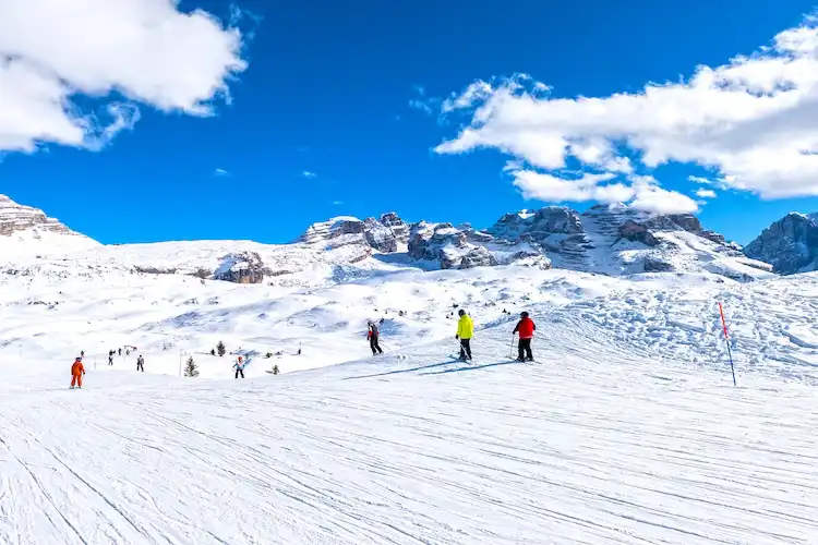 Pessoas praticando ski em pista de neve ampla, com montanhas cobertas de neve ao fundo e céu azul.