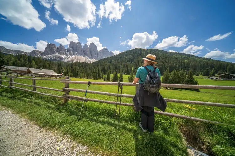 Pessoa observa as montanhas de Val di Funes, nas Dolomitas, em meio a um campo verde sob céu azul.