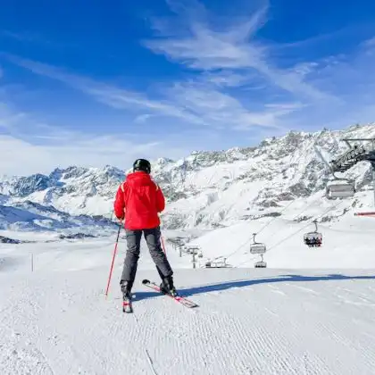 Pessoa praticando ski em pista de neve, com montanhas cobertas de gelo ao fundo e teleféricos ao redor.