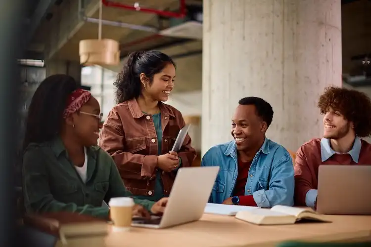 Estudantes interagem em mesa de estudos com laptops e materiais.