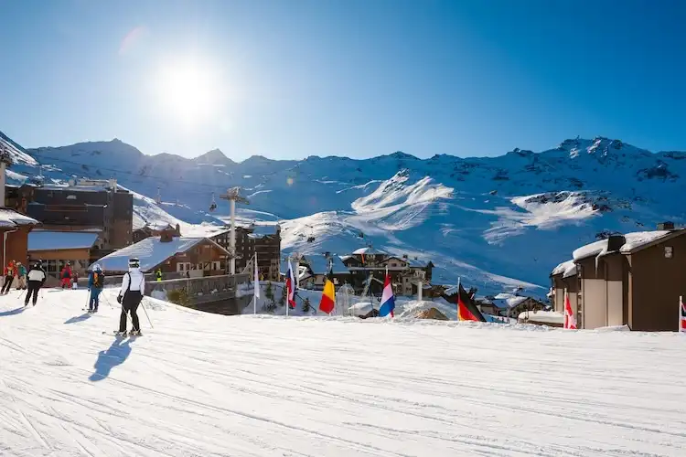 Pessoas praticam esqui em estação de inverno com montanhas nevadas ao fundo.