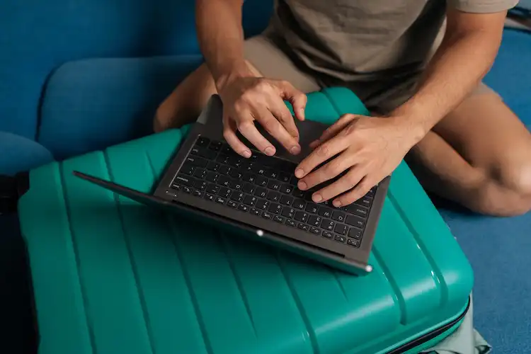 Pessoa utiliza notebook apoiado sobre mala de viagem.